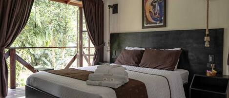 A bedroom with a large bed, dark curtains, a painting on the wall, and a nightstand with a lamp.