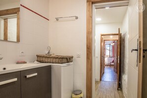 Casa de banho com lavatório branco, armários de madeira, cesto de vime e porta visível para outro espaço.