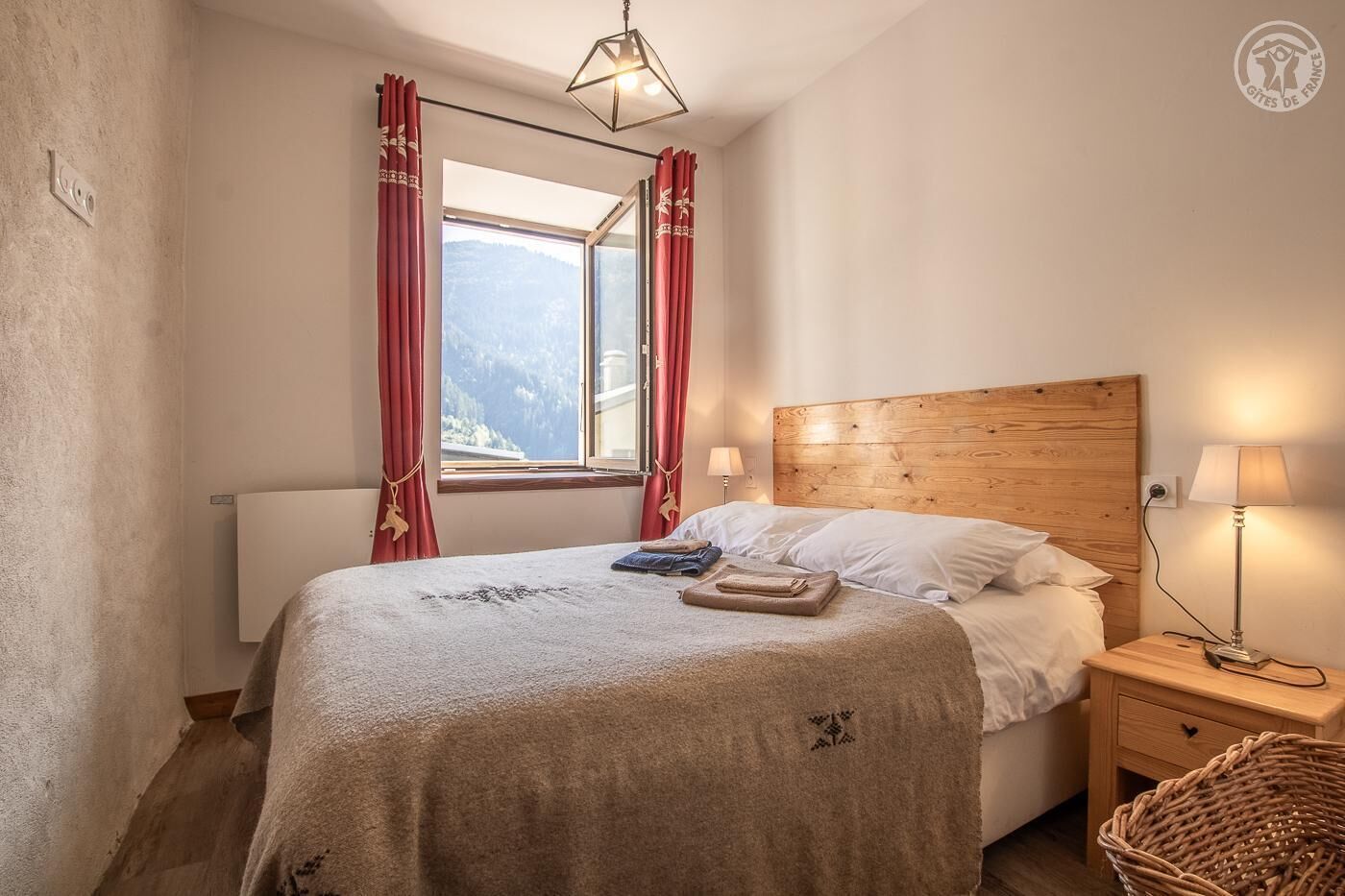 A bedroom with a wooden headboard, a bed with white linens, a window with red curtains, a bedside lamp, and a wicker basket.