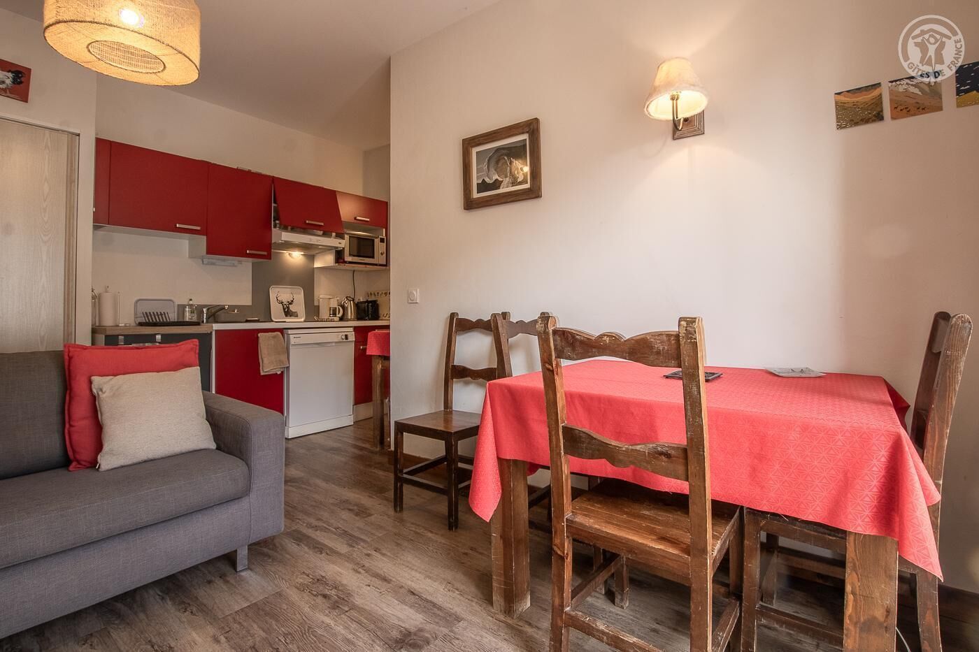 Un espacio habitable compacto con kitchenette, una zona de comedor con mesa roja y sillas de madera, y un sofá gris con cojines rojos.