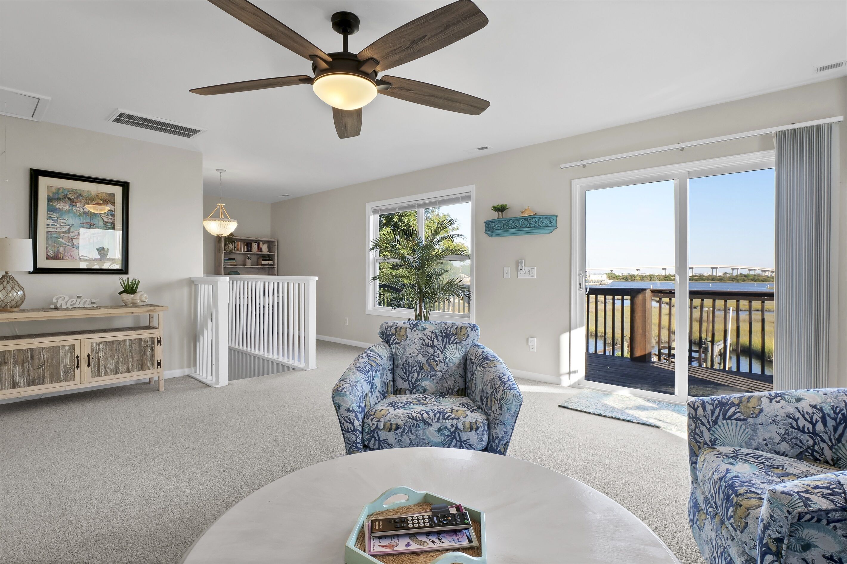 A spacious living room with a ceiling fan, a large window with a view of the outdoors, a white entertainment unit, and a patterned armchair.