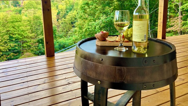 Una mesa de madera con una botella de vino, una copa y un cuenco de fruta en una terraza con vista a un paisaje boscoso.