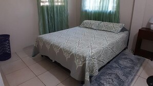 A bedroom with a bed, green curtains, a patterned bedspread, a blue rug, and a small nightstand.