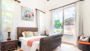 A bedroom with a large bed, a nightstand, a wicker bench, and a window with curtains.