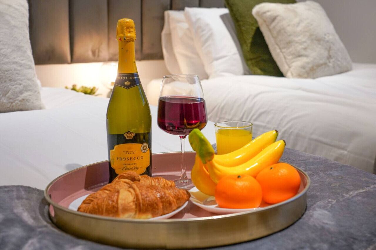 Bandeja de desayuno con una botella de Prèsecco, una copa de vino tinto, un croissant, plátanos y naranjas sobre una cama con ropa blanca y almohadas.