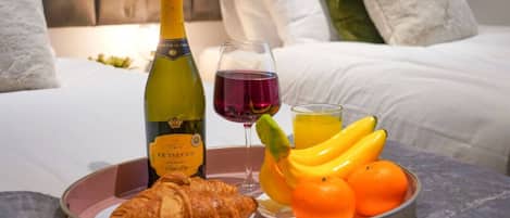 A breakfast tray with a bottle of Prèsecco, a glass of red wine, a croissant, bananas, and oranges on a bed with white bedding and pillows.