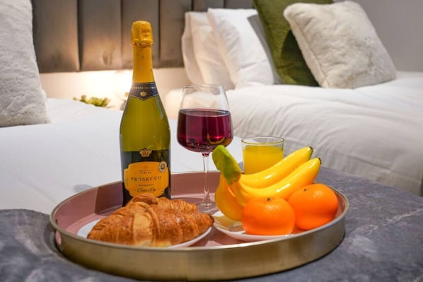 A breakfast tray with a bottle of Prèsecco, a glass of red wine, a croissant, bananas, and oranges on a bed with white bedding and pillows.