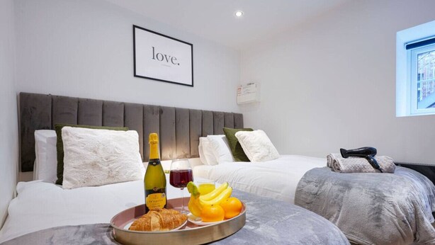 A hotel room with two beds, a framed word 'love' on the wall, a tray with croissants, fruit, and a bottle of wine.