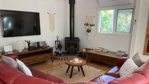 A cozy living room with a red leather sofa, a flat-screen TV, a fireplace, and a window with a view of greenery. - Historic Cottage 4 Riverside Dve. Pet friendly, flat fenced yard  on the River. (Warburton)