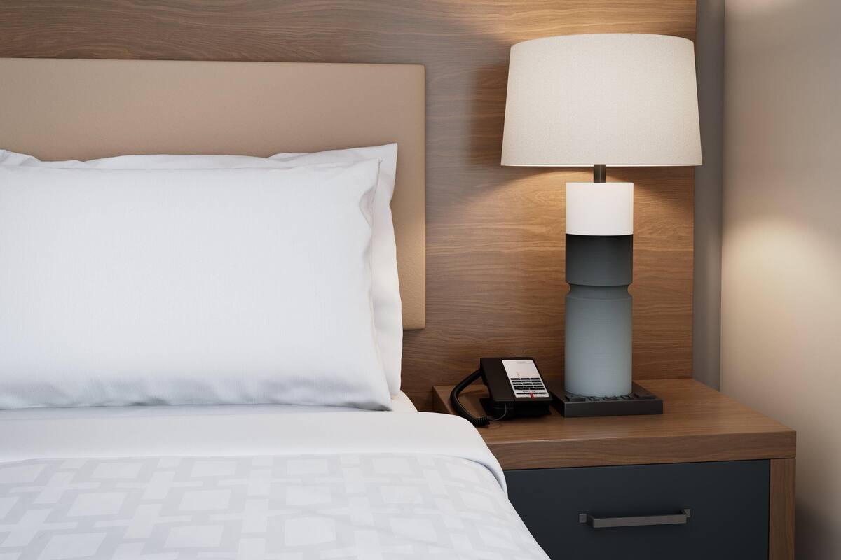 Ein ordentlich gemachtes Bett mit einem weißen Kopfkissen und einer grauen, gemusterten Bettdecke, ein Nachttisch mit Lampe und Telefon sowie eine Schublade darunter.