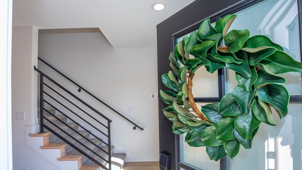 Un interior moderno con una escalera, una puerta adornada con un adorno navideño verde y una ventana con persianas.