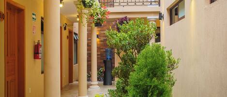 Pasillo de hotel con columnas, un arbusto verde en el centro y macetas colgantes con flores.