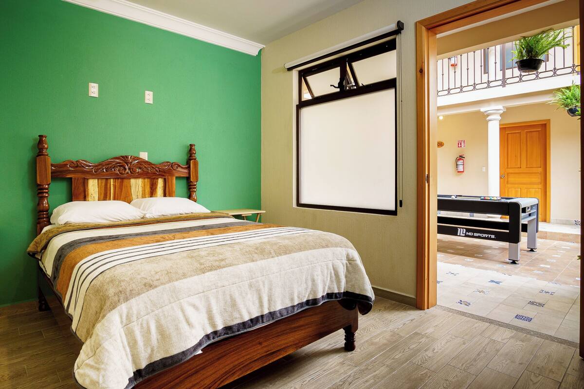 Un lit bien fait, avec une tête de lit en bois, une grande fenêtre donnant sur l’extérieur et une table de billard dans la pièce adjacente.