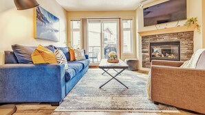 A living room with a blue sofa, a brown armchair, a fireplace, and a television mounted above it. - Cozy 2 Bedroom/3 Beds/Mountain View /AC/Free Parking (Canmore)
