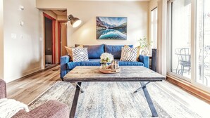 A living room with a blue sofa, a wooden coffee table with a decorative tray, and a large window with a view of the city. - Cozy 2 Bedroom/3 Beds/Mountain View /AC/Free Parking (Canmore)