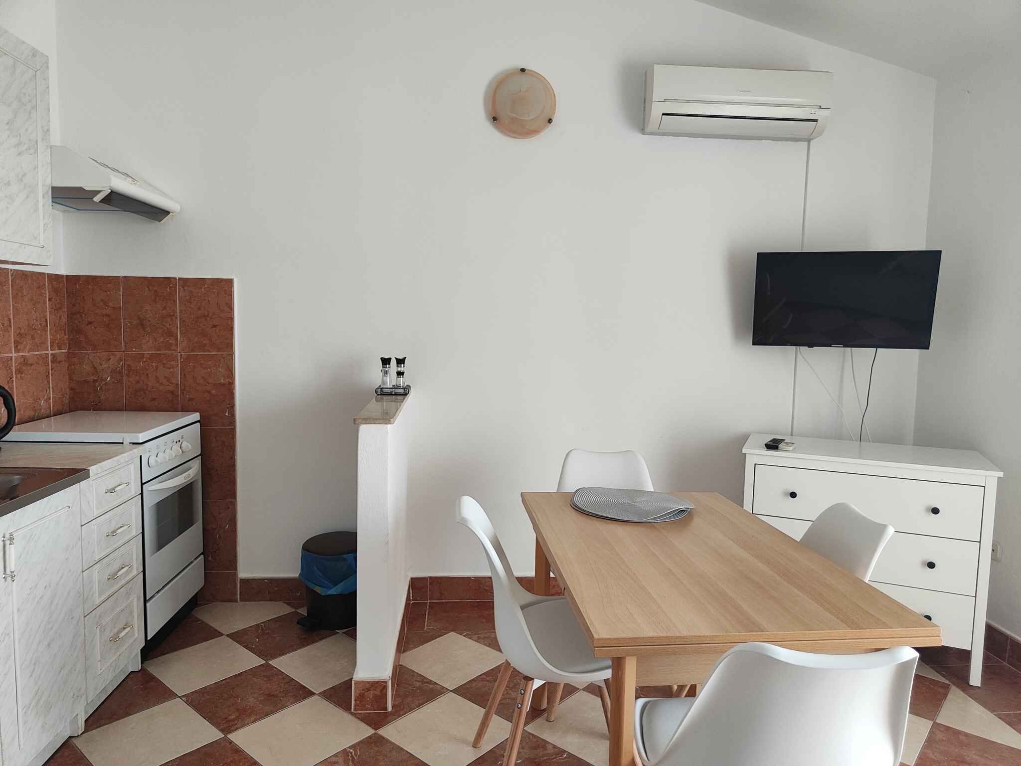 A compact kitchen with a stove, oven, and white cabinets. A dining table with chairs is in the center, and a TV is mounted on the wall.