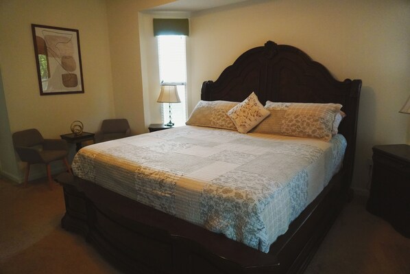 A neatly made bed with a floral bedspread and two pillows, a wooden headboard, a small bedside table with a lamp, and a chair in a room with beige walls and a framed picture on the wall. - Home Feel with a Vacation Ambience (Port St. Lucie)