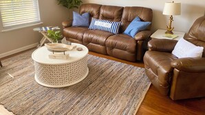 A brown leather sofa with blue and white striped pillows, a round coffee table with decorative items, a striped rug, and a painting of a boat on water.