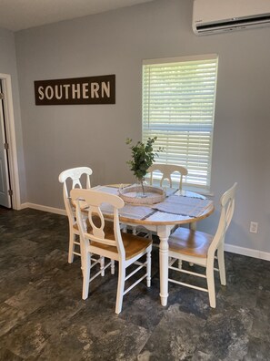 A dining area with a round table, four white chairs, a potted plant, and a sign on the wall.