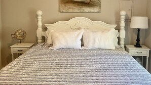 A bedroom with a patterned bedspread, a white headboard, a wall-mounted painting of a boat, a bedside lamp, and a small decorative fish sculpture.