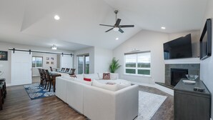 A spacious living room with a large white sofa, a flat-screen TV mounted above a fireplace, and a dining area with a wooden table and chairs. - Mountain Views, Single family home near airport. Skyview Retreat! (Belgrade)
