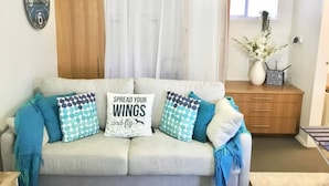 Una sala de estar con sofá, almohadas, un reloj, una ventana con cortinas, un mueble de madera y un jarrón con flores.