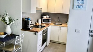 Cocina compacta con microondas, tostadora y lavabo. Pequeño comedor con una silla y un jarrón de flores.
