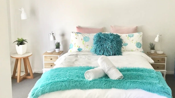 Un dormitorio ordenado con cama, almohadas, una manta azul y un cuadro en la pared.
