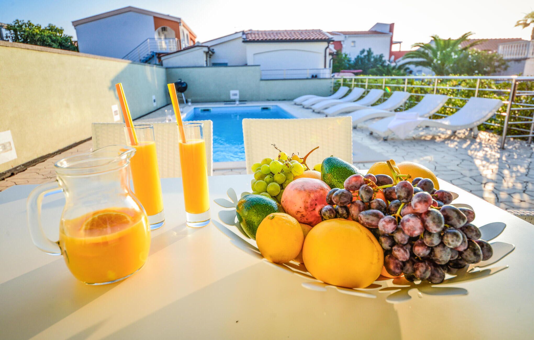 A table with a fruit platter and two glasses of orange juice, a pool, and lounge chairs in the background.