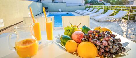 A table with a fruit platter and two glasses of orange juice, a pool, and lounge chairs in the background.