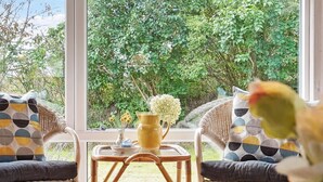 Een rieten stoel met een kussen, een klein tafeltje met een gele vaas en een glas sinaasappelsap, en uitzicht op een weelderige tuin door grote ramen.