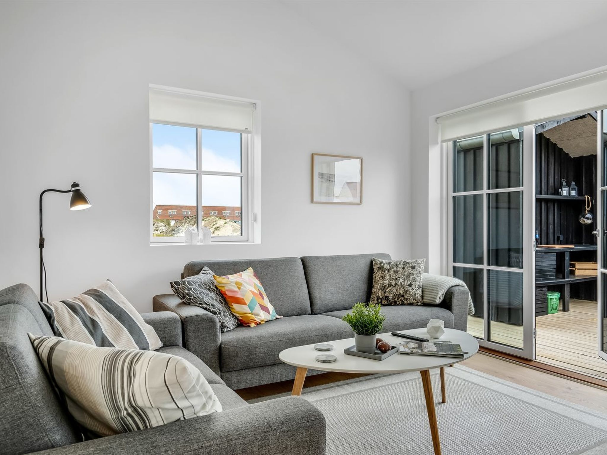 Un salon moderne avec un canapé gris, une petite table basse, une lampe sur pied et une grande porte-fenêtre coulissante donnant sur un espace extérieur.