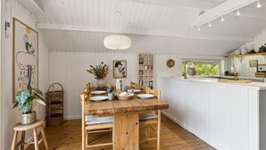 Un comedor con mesa y sillas de madera, una cocina blanca con pileta y estanterías, y un techo con vigas de madera a la vista.