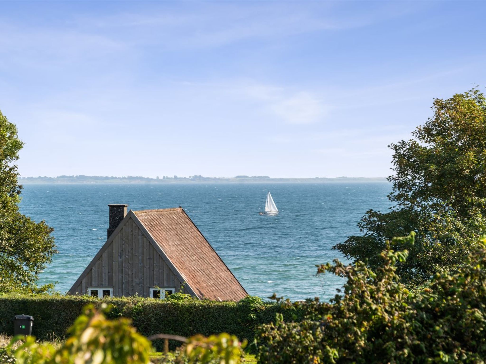 Een houten huis met schuin dak dat uitkijkt over een rustige zee met een zeilboot.