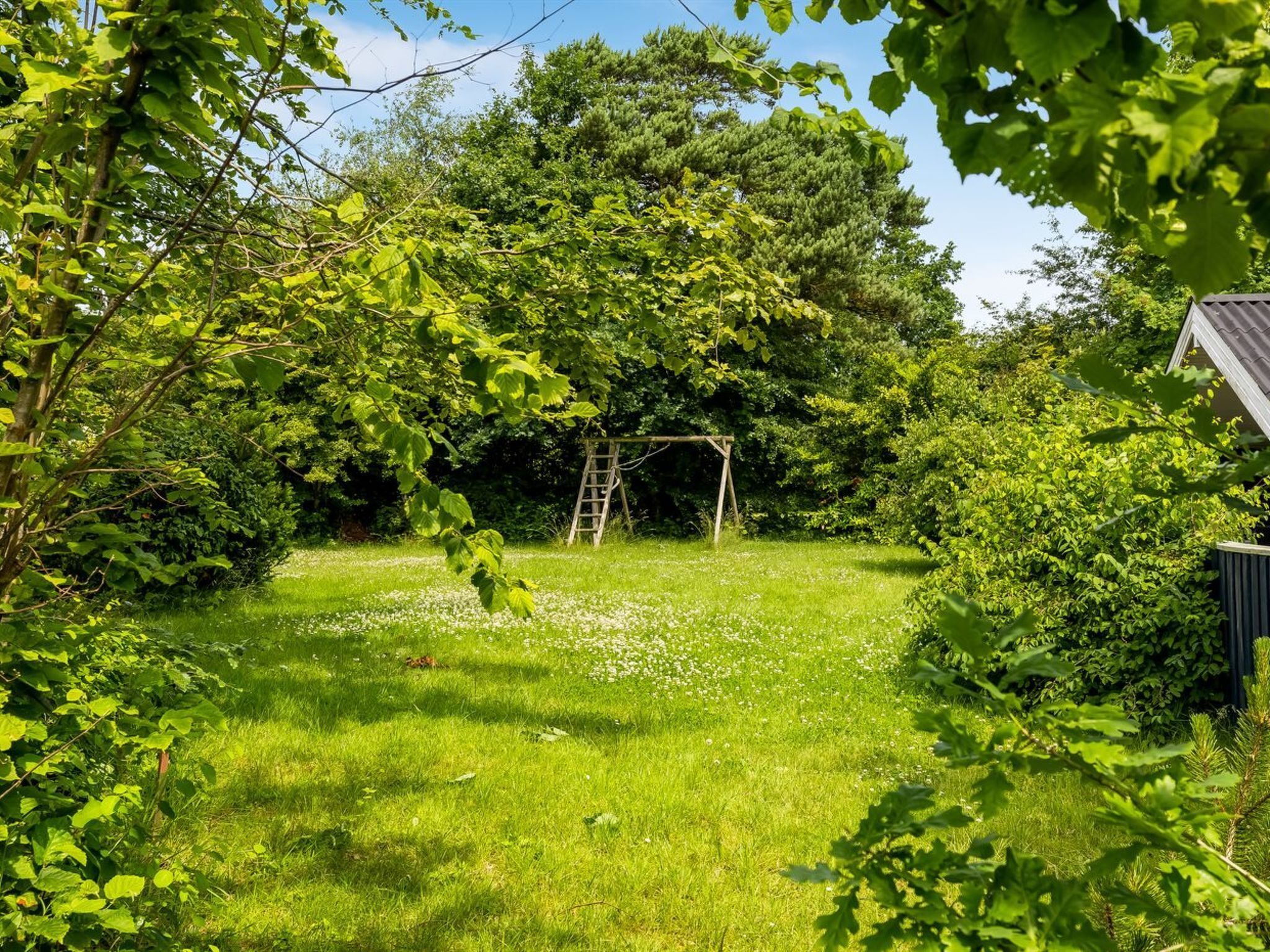 A lush garden with a wooden swing set, surrounded by dense foliage and a clear sky.