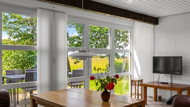Ein Zimmer mit Holztisch, Fernseher und großen Fenstern mit Blick ins Grüne.