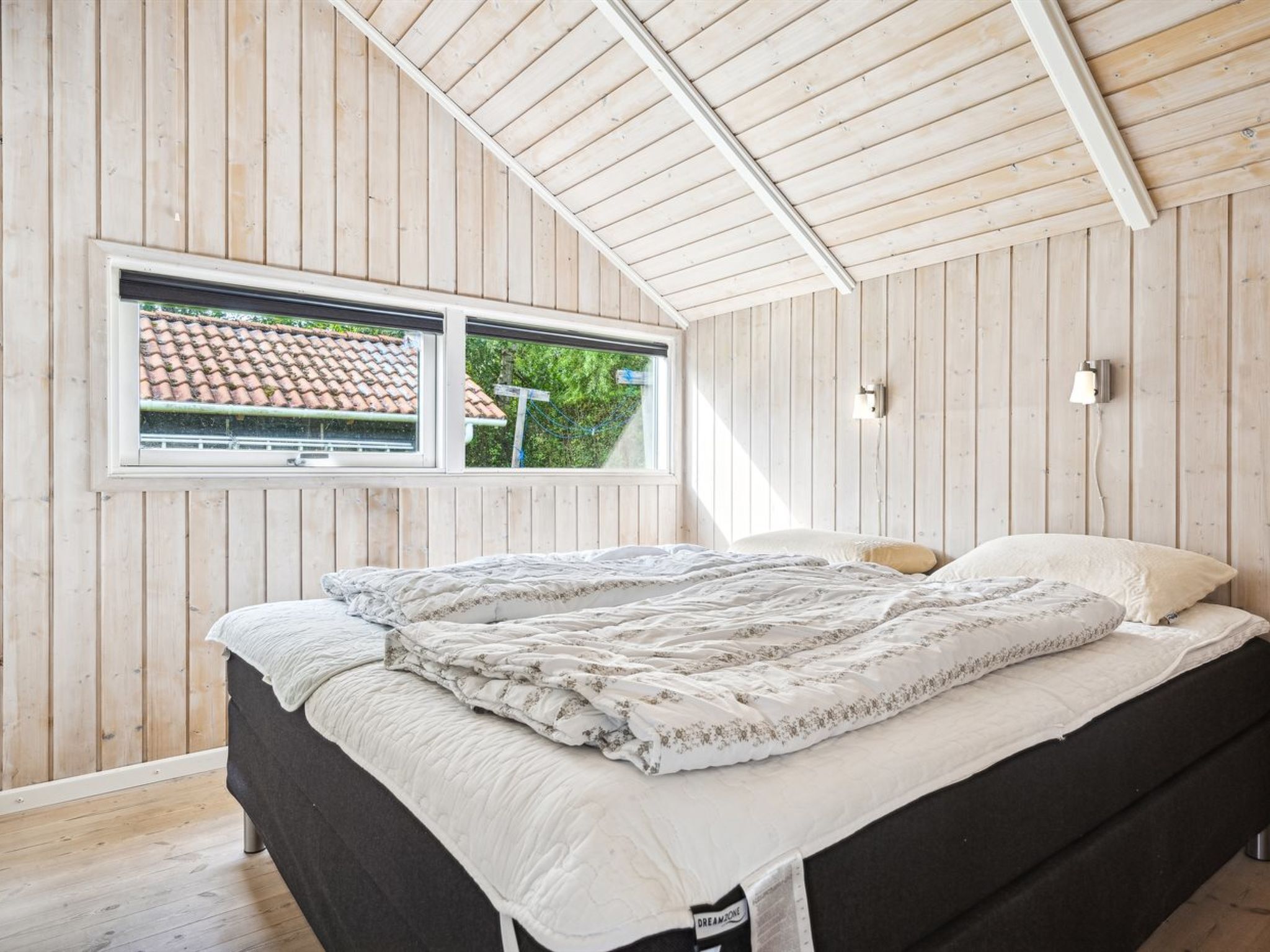 Une chambre avec un plafond en pente, des murs en bois, un lit recouvert d’une courtepointe, une fenêtre donnant sur une maison et un luminaire fixé au mur.