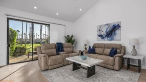 A modern living room with two beige sofas, a marble coffee table, and a view of a landscaped garden through large sliding glass doors. - Luxury Palm Valley 3BR  3 bath condo (Palm Desert)