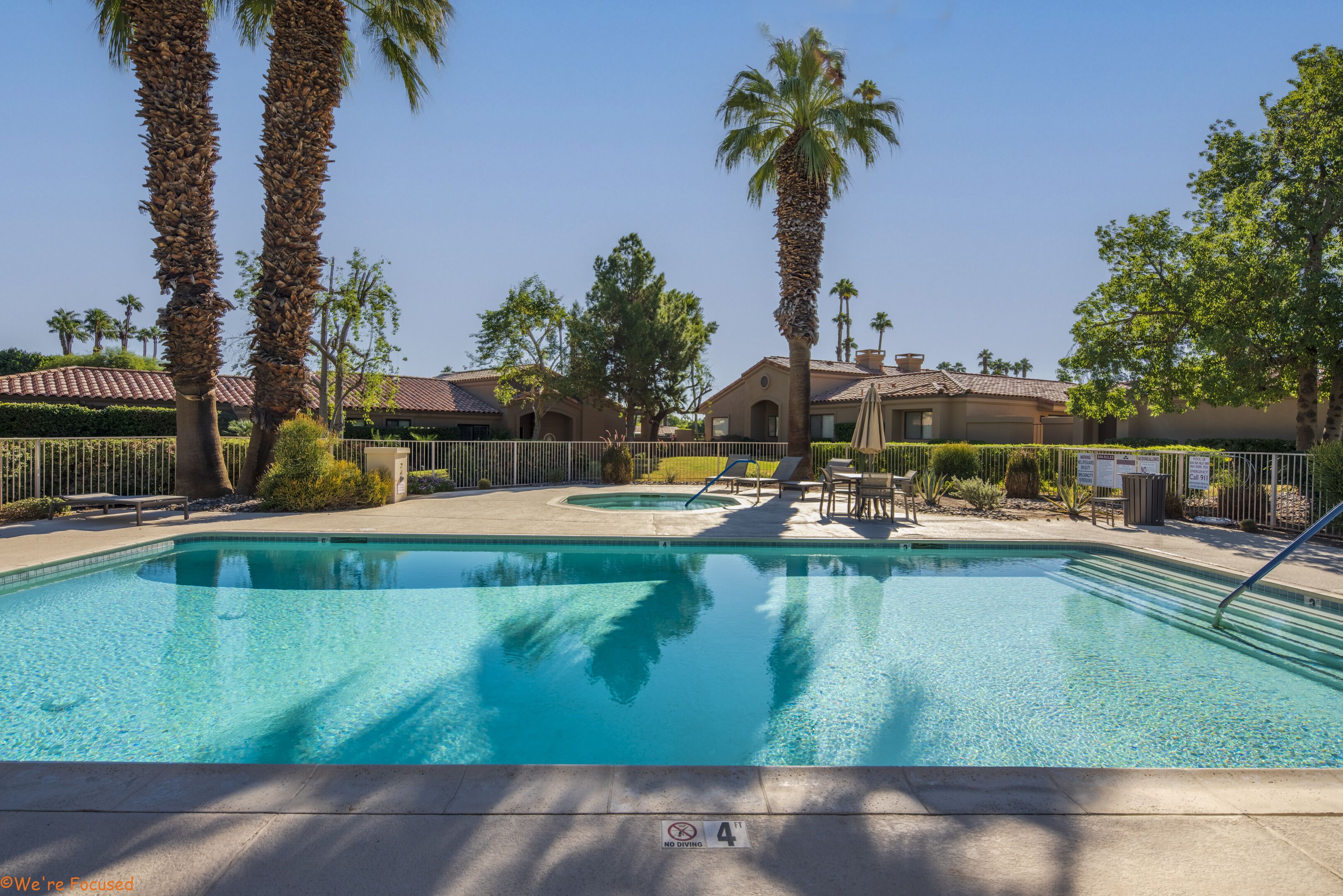 A swimming pool with palm trees, a lounge area, and a building with a tiled roof in the background.