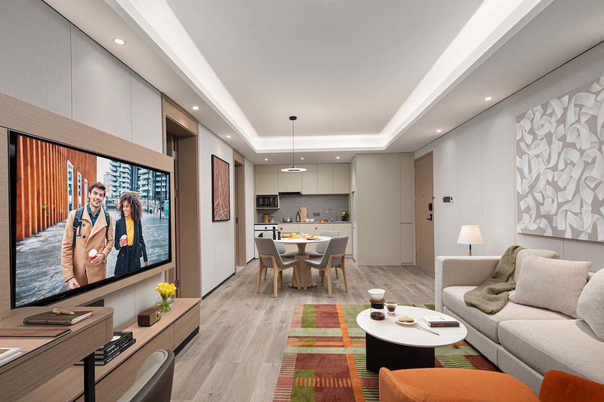 Een moderne woonkamer met een flatscreen televisie, een eethoek met een ronde tafel en stoelen, een bank en een keuken op de achtergrond.