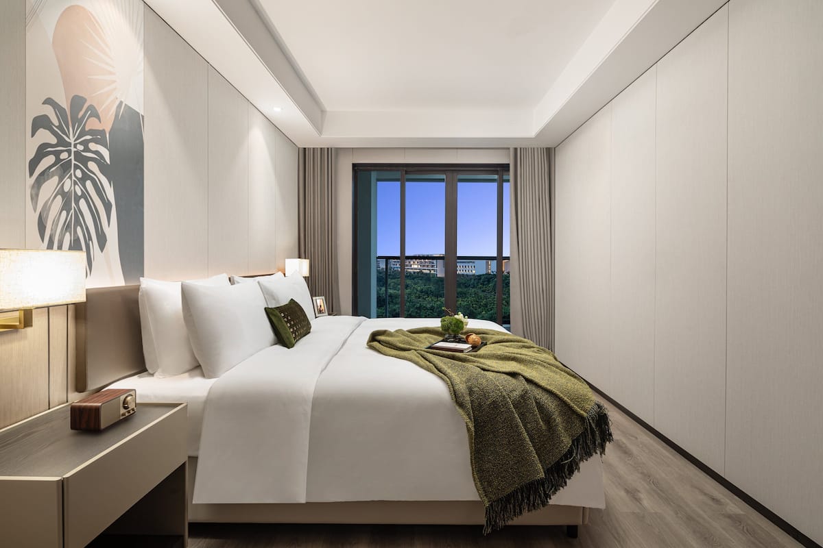 Een moderne hotelkamer met een groot bed, een nachtkastje, een lamp en uitzicht op de stad door grote ramen.