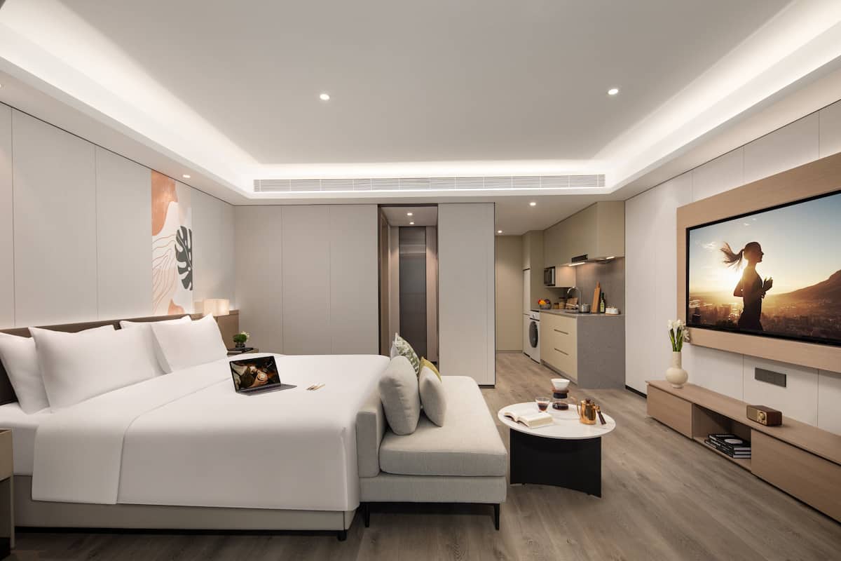 Een moderne hotelkamer met een groot bed, een kleine zithoek, een salontafel, een televisie en een kitchenette op de achtergrond.