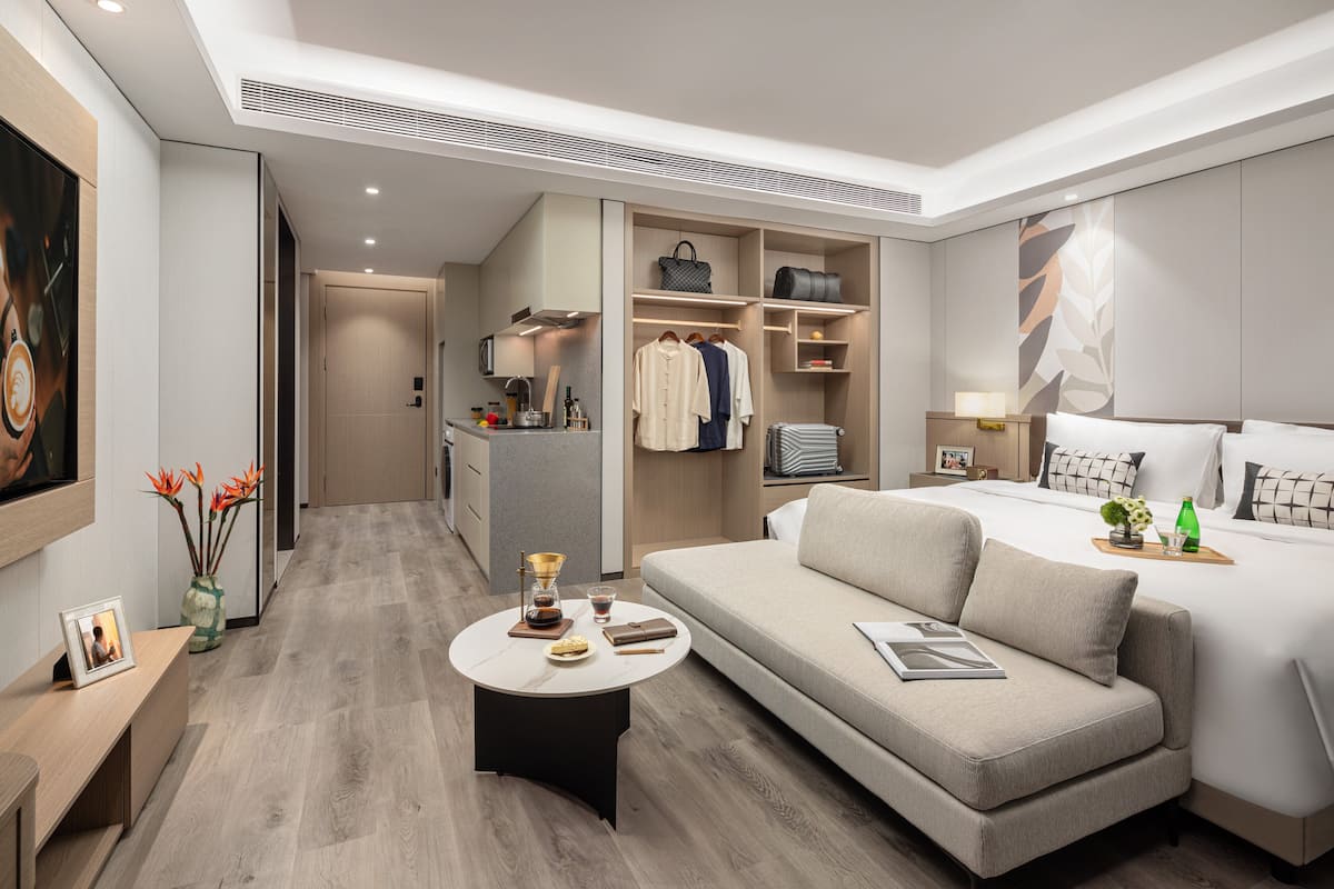 Een moderne hotelkamer met een bank, nachtkastje, lamp, bed, garderobe en een klein keukenhoekje.