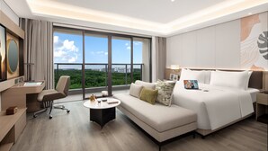 A modern hotel room with a large bed, a desk, a chair, a coffee table, and a view of the city. - Citadines Hope City Boao (Qionghai)