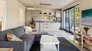 A modern living space with a grey sectional sofa, a white coffee table, and a kitchen area with white cabinets and a large window. - "Uma" - 1.5km from the sea by Interhome (Knebel)
