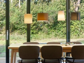 Un espace repas avec une longue table et des chaises, de grandes fenêtres ornées de lampes suspendues, et une vue sur la verdure à l’extérieur.