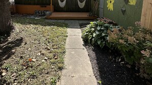 A residential backyard with a concrete pathway, a wooden fence, and a house with a green exterior. - Charming 1920’s shotgun in southern Indiana, minutes from downtown Louisville  (New Albany)