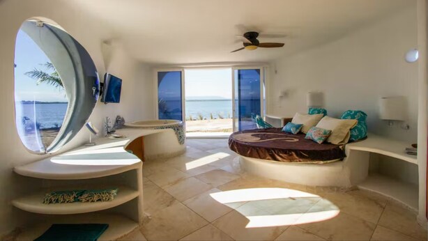 A bedroom with a large window, a bed, a ceiling fan, and a view of the sea. - Lion Fish Retreat (Placencia)