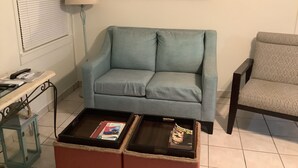 Uma sala de estar com um sofá azul-clarinho, uma poltrona estampada, uma mesa de centro com revistas, um abajur e um espelho com moldura de concha.