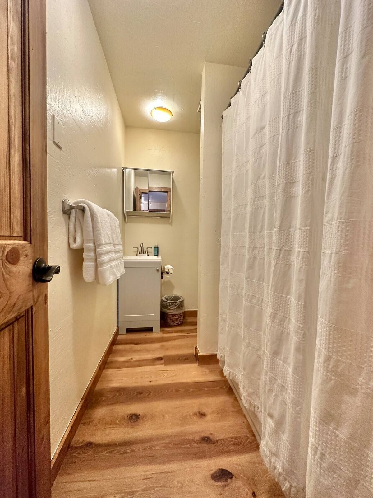 Ein schmaler Flur mit Holzboden, der zu einem Badezimmer führt. Ein weißer Vorhang verdeckt den Duschbereich, und ein kleines Spiegelbild hängt an der Wand.